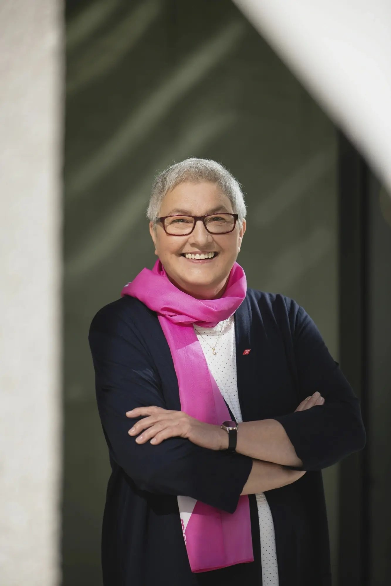 Lächelnde Frau (Elke Hannack) mit Brille und pinkem Schal vor neutralem Hintergrund, mit verschränkten Armen.