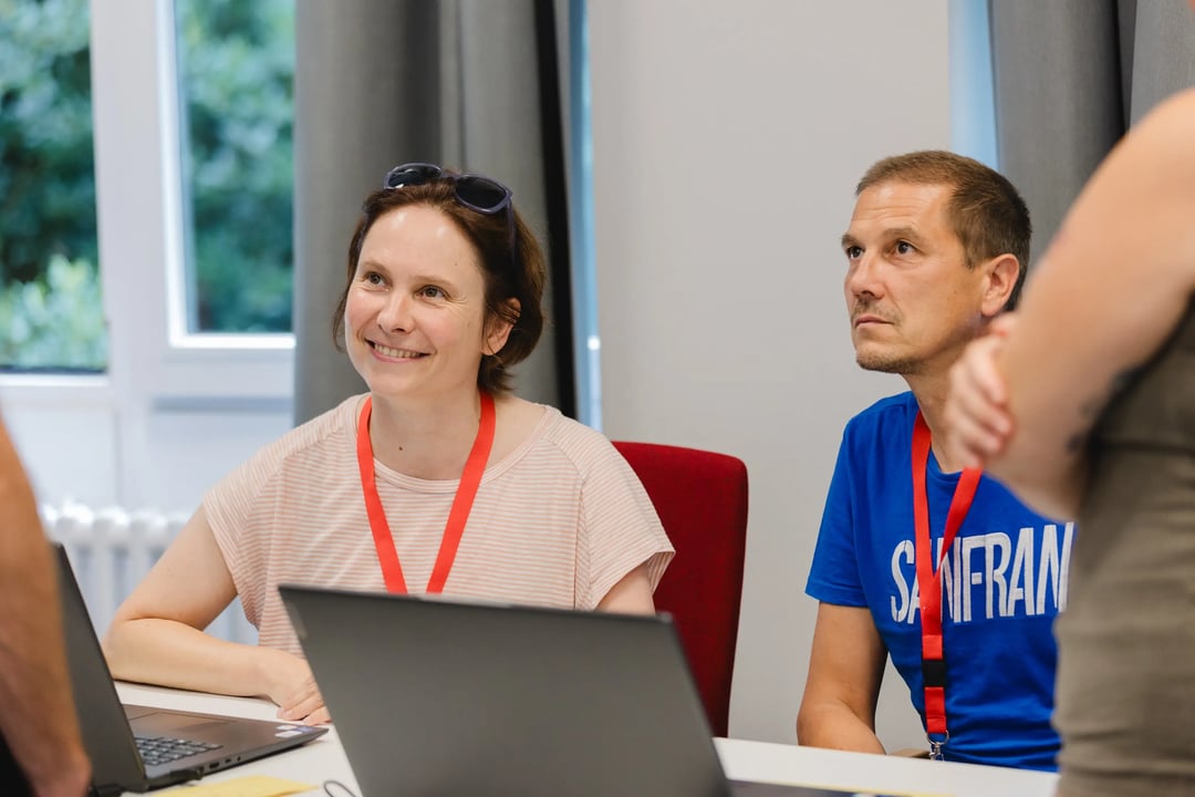 Zwei Personen sitzen an einem Tisch mit Laptops und roten Schlüsselbändern um den Hals, im Hintergrund graue Vorhänge und ein Fenster mit Blick ins Grüne.