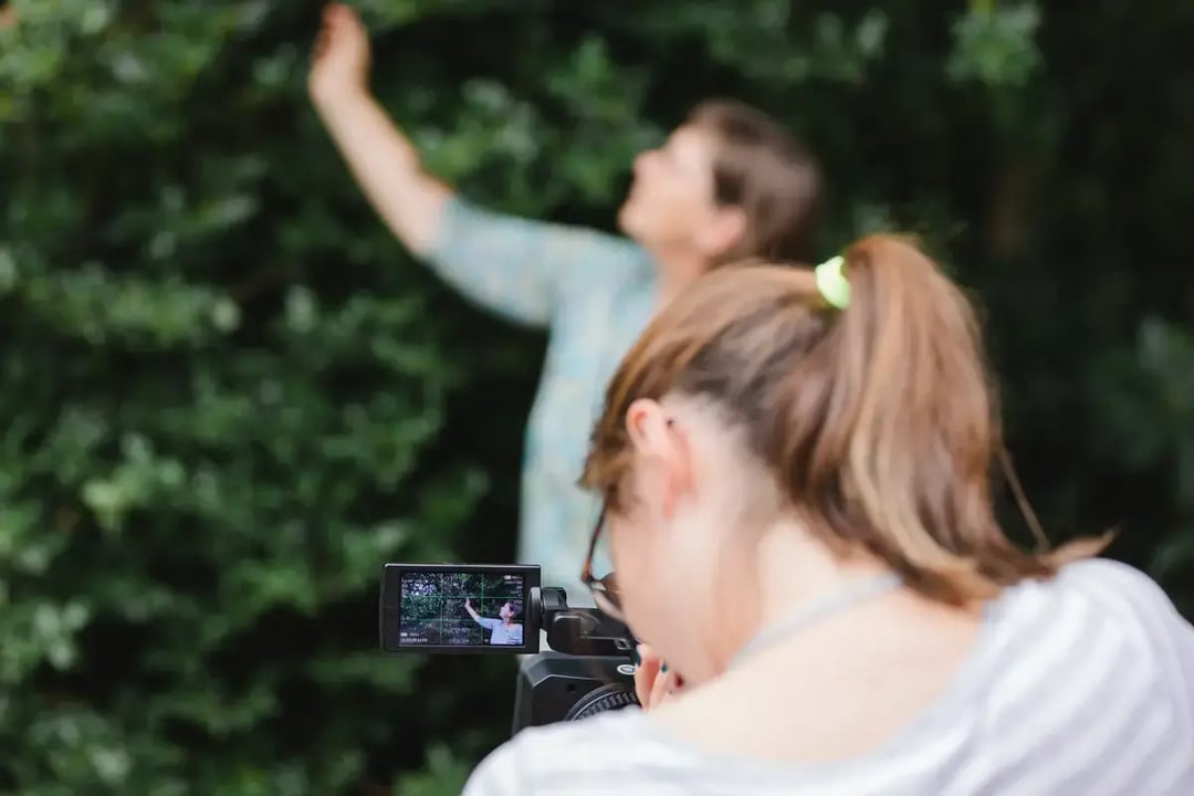 Person filmt mit einer Videokamera eine andere Person vor grünem Buschwerk, Kameradisplay zeigt die Aufnahme.