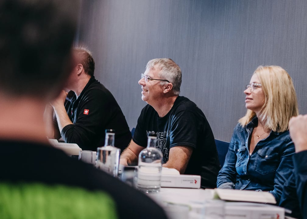  Seminarteilnehmende sitzen an einem Tisch mit Wasserflaschen und Unterlagen; graue Wand im Hintergrund.