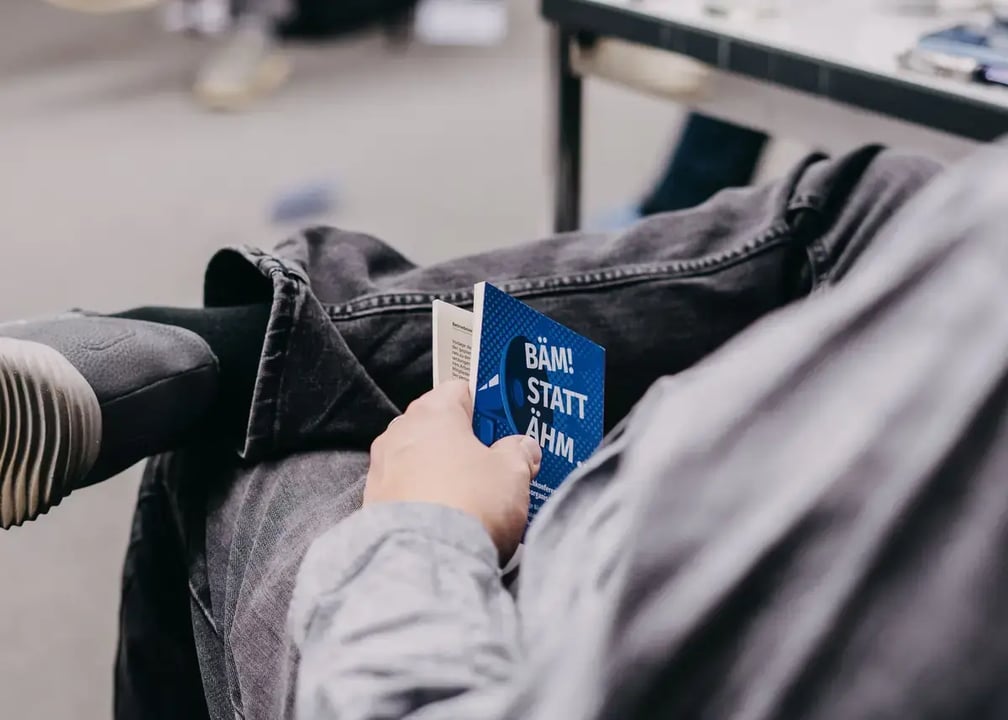 Person hält ein kleines blaues Buch mit der Aufschrift „BÄM! STATT ÄHM“ in der Hand, sitzt mit überkreuzten Beinen neben einem Tisch.