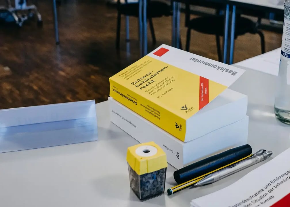 Auf einem Tisch liegen ein dicker gelber Gesetzeskommentar, ein weißes Buch, ein Bleistift, zwei Stifte und ein gelber Spitzer. Im Hintergrund stehen Tische und Stühle eines Seminarraums.