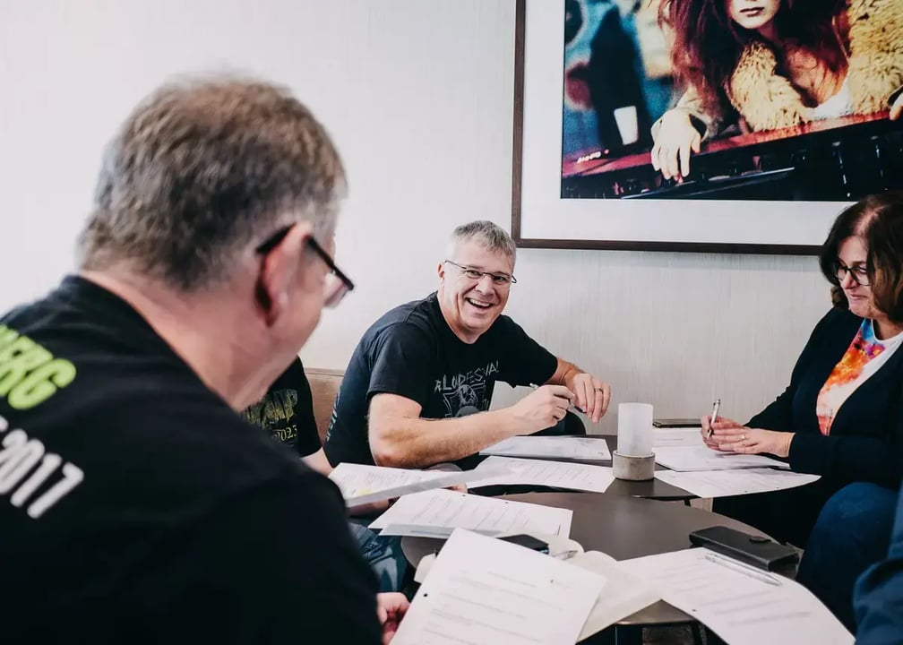 Gruppe von Personen sitzt an einem Tisch mit mehreren Dokumenten und Stiften; im Hintergrund hängt ein großes gerahmtes Bild.