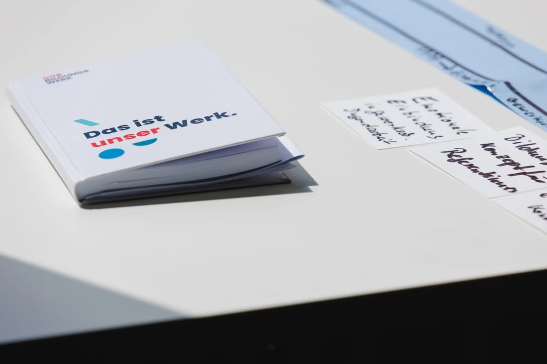 Weißes Heft mit der Aufschrift „Das ist unser Werk.“ liegt auf einem Tisch neben handgeschriebenen Notizzetteln.