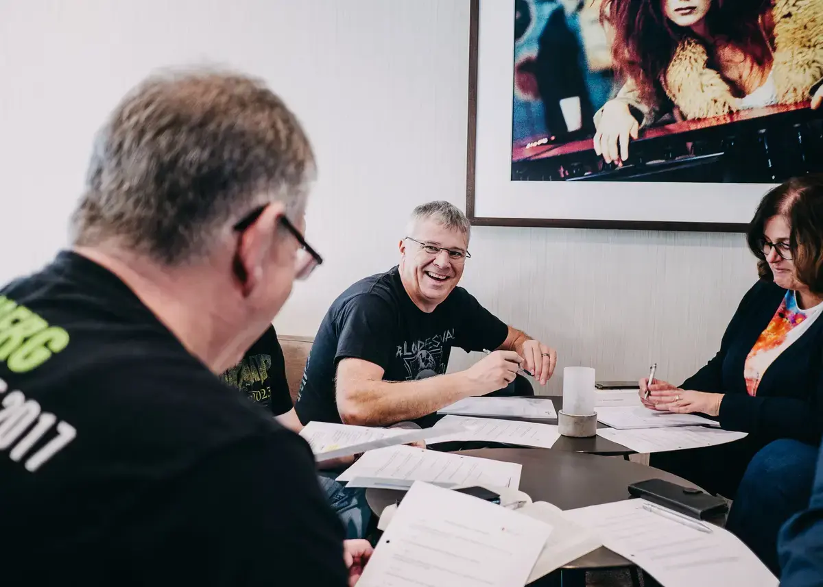 Gruppe von Personen sitzt an einem Tisch mit mehreren Dokumenten und Stiften; im Hintergrund hängt ein großes gerahmtes Bild.